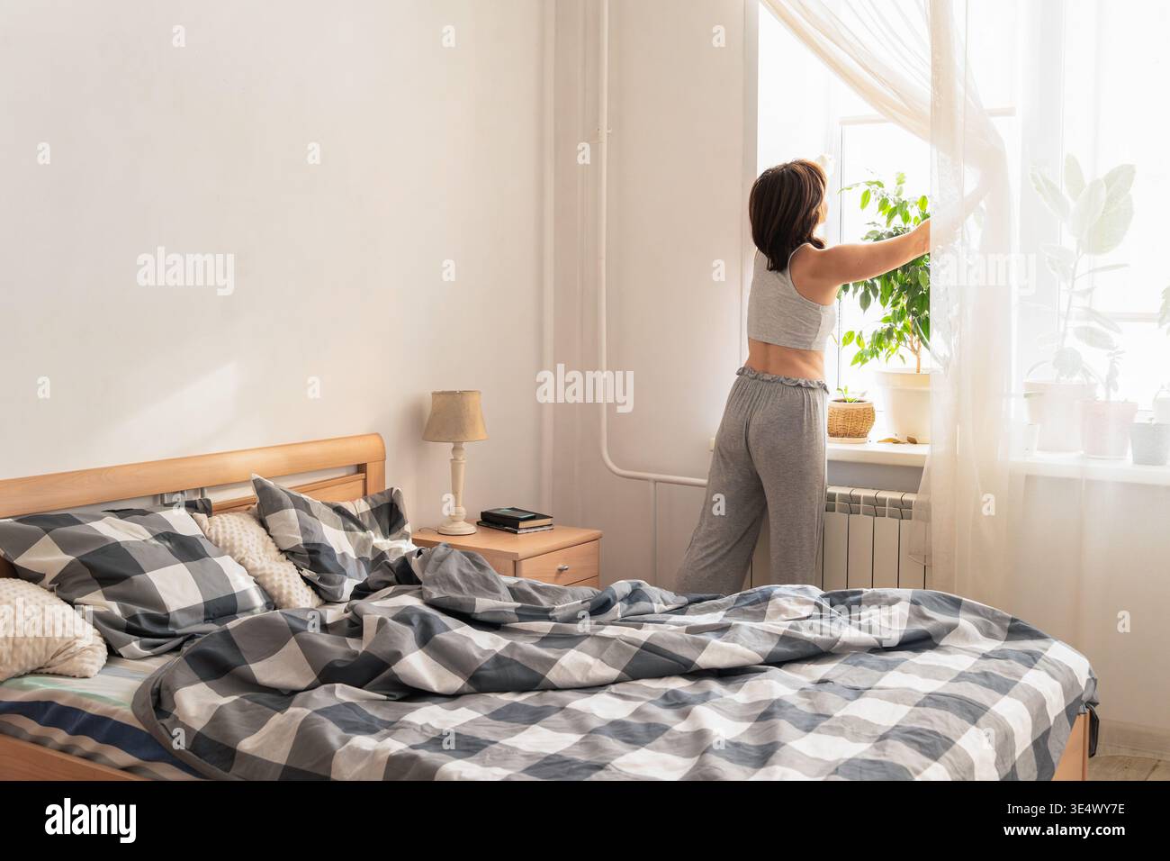 Frau im Pyjama öffnet Vorhänge, begrüßt Morgensonne in ihrem Schlafzimmer und beginnt den Tag mit einer Wellness-Routine. Authentischer Lifestyle Stockfoto