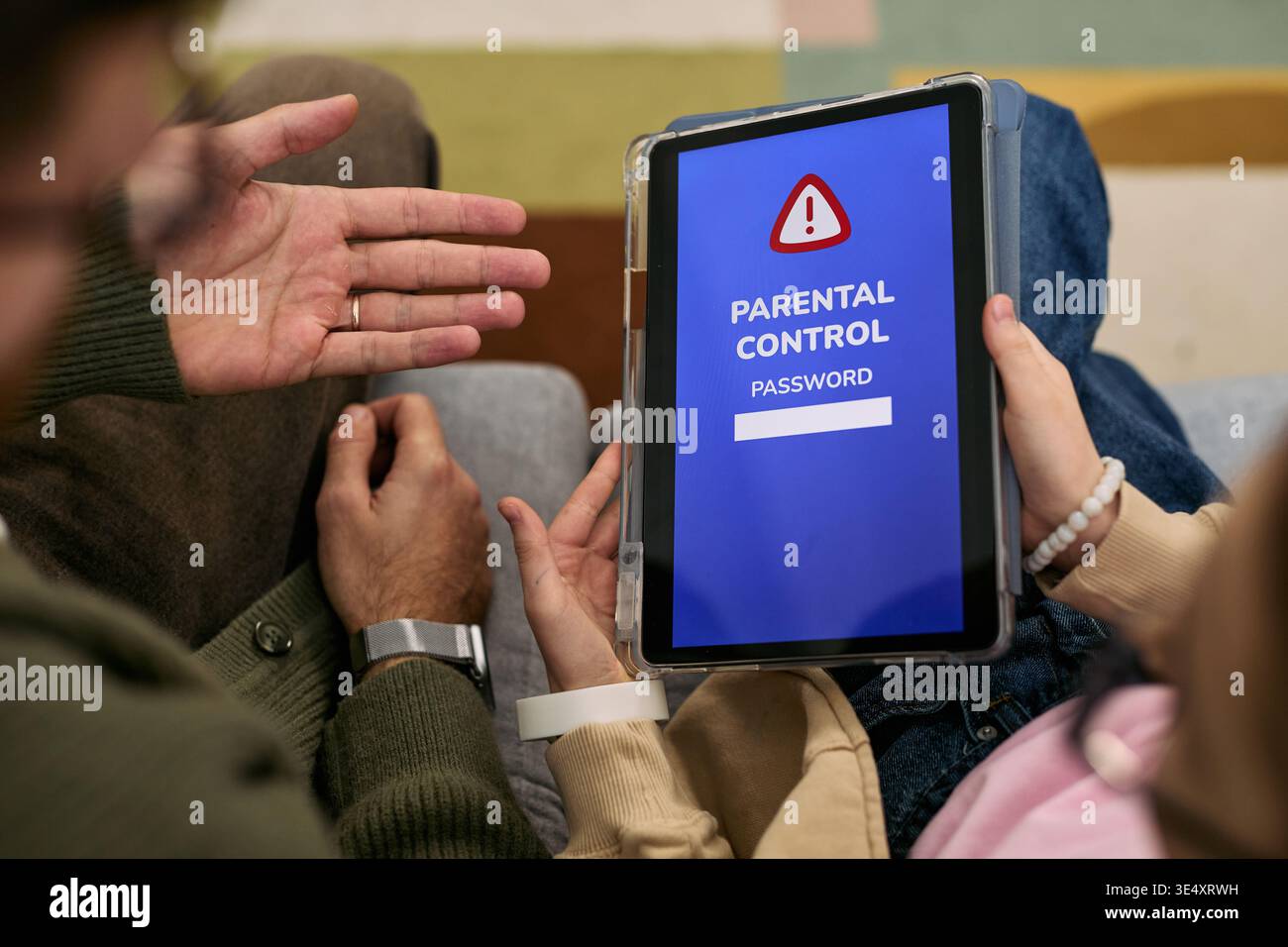Kaukasischer Mann mittleren Alters, der Gesten gibt, während ein kaukasisches Kind ein digitales Tablet hält, das das Passwort der Elternkontrolle anzeigt, beide sitzen eng zusammen auf dem Boden, Hände sichtbar Stockfoto