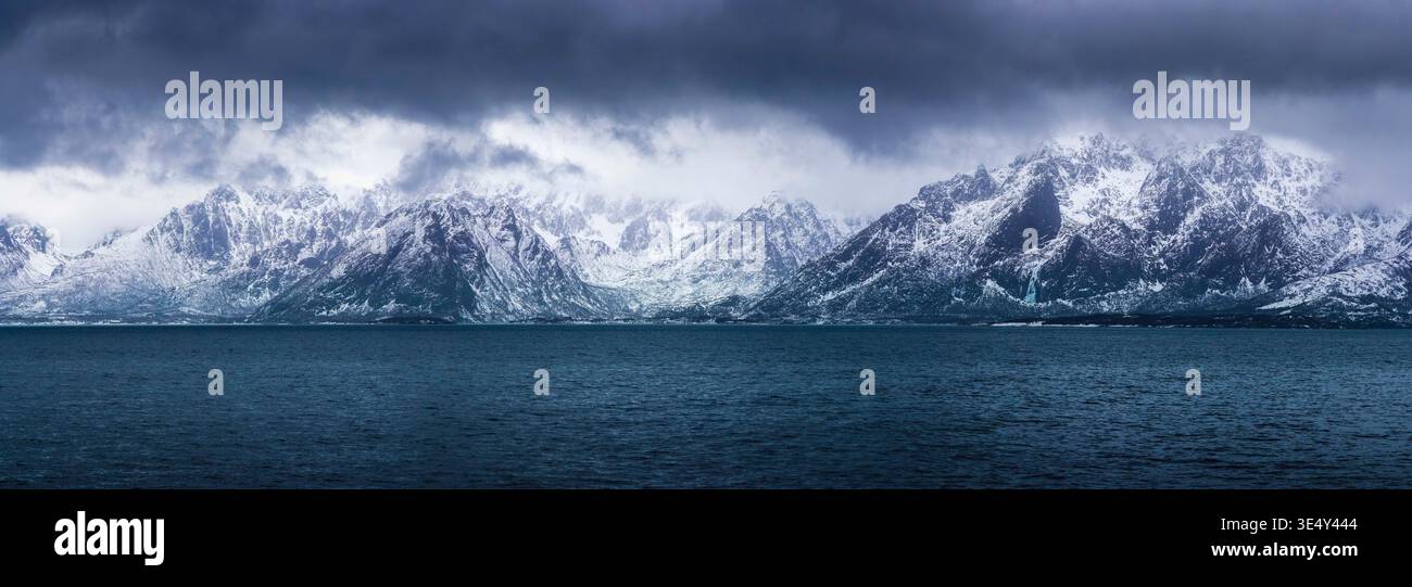 Gefrorene Gipfel erheben sich dramatisch, in weiß getarnt, und erstrecken sich über den Himmel. Das dunkle Wasser reflektiert die düsteren Wolken und schafft einen markanten Kontrast. Stockfoto