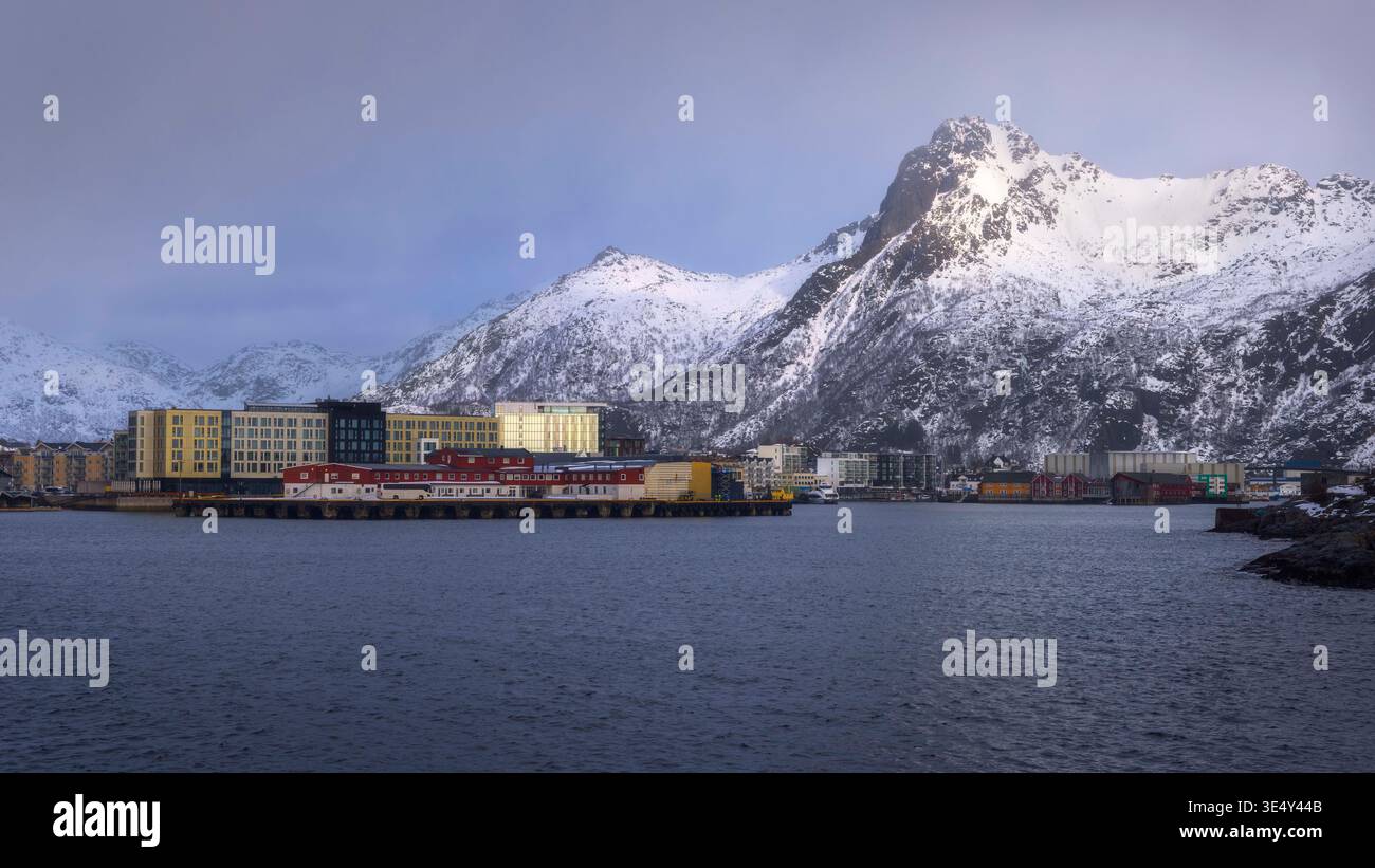Im ruhigen Hafen von Svolvaer, Lofoten, säumen farbenfrohe Gebäude die Küste unter einer Decke aus weichen Wolken. Schneebedeckte Berge überragen majestätisch Stockfoto