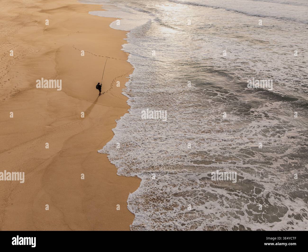Aus der Vogelperspektive steht eine Einzelfigur auf dem braunen Sand des Strandes, während die Wellen hereinbrechen und lange Schatten werfen, Portugal. Stockfoto