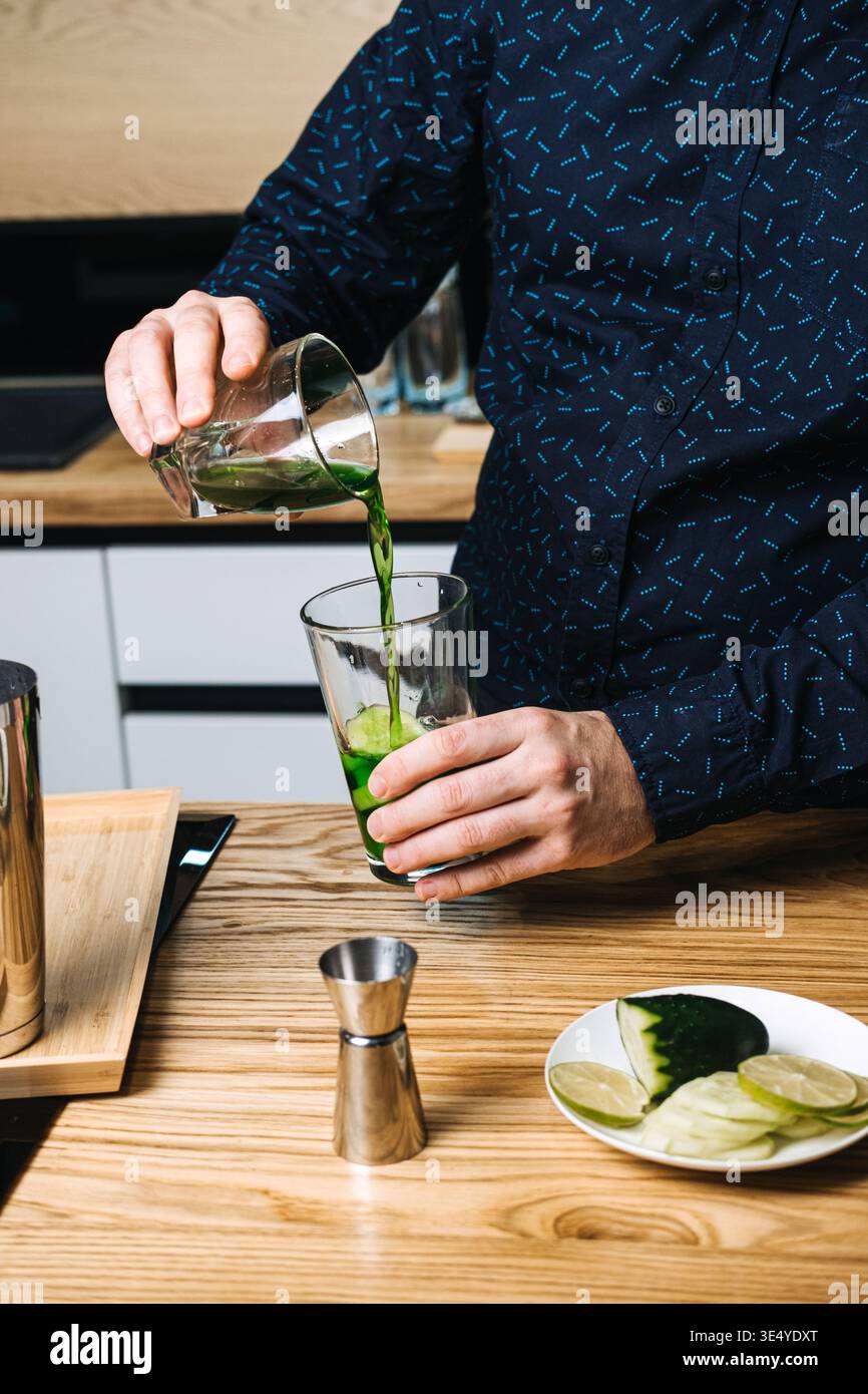 Hände halten Glas der grünen Flüssigkeit über das Mischglas mit Gurkenscheiben auf Holztheke. Chlorophyllwasser-Vorteile, funktionelle grüne Getränke Stockfoto