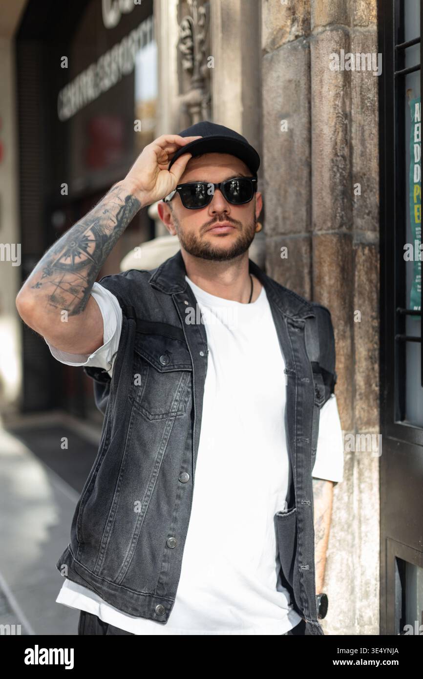 Ein cooler, gutaussehender Mann in modischer Kleidung mit Sonnenbrille trägt eine schwarze Kappe und spaziert durch Barcelona Stockfoto