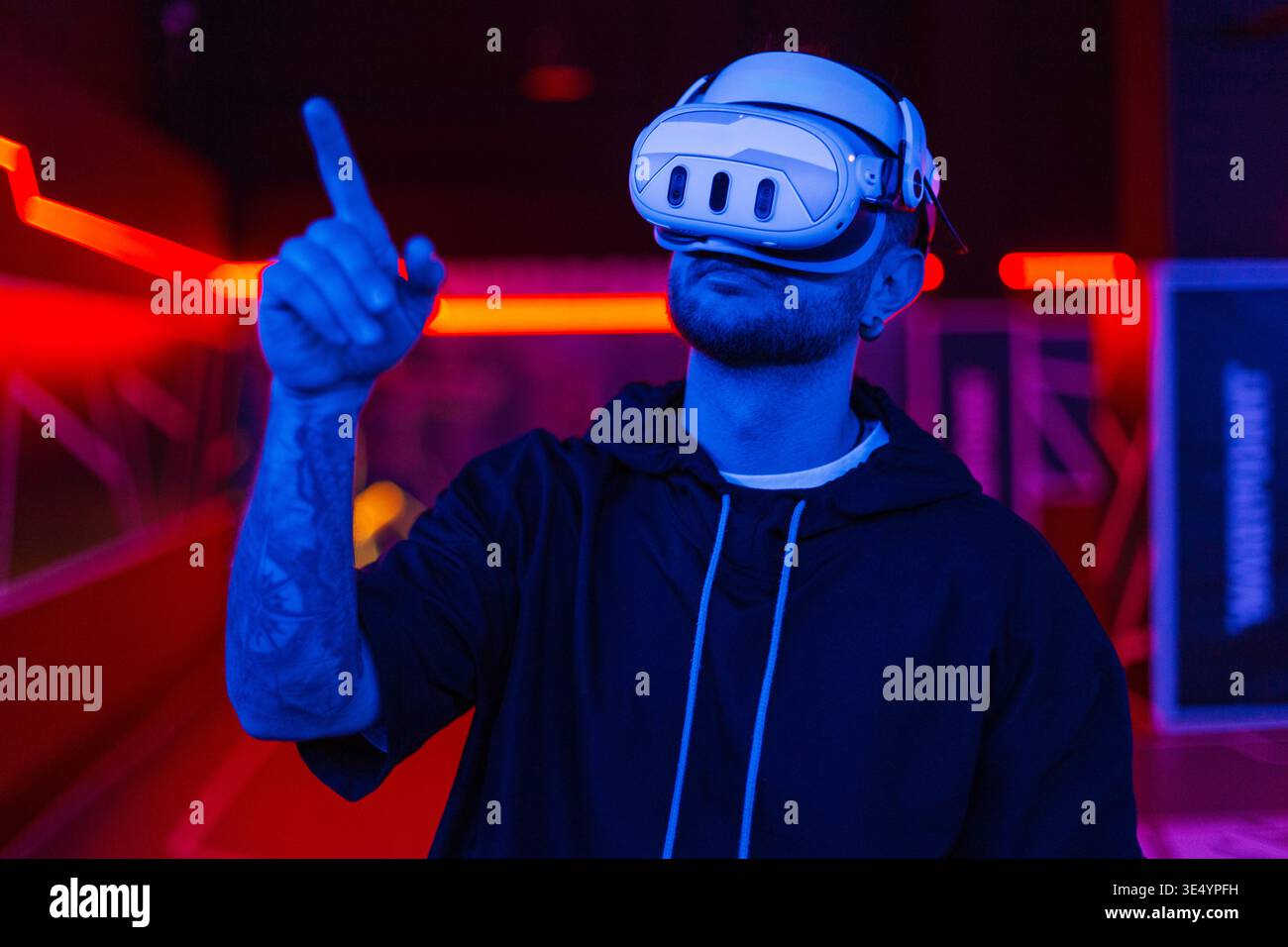 Hübscher Mann mit VR-Brille in einem schwarzen Hoodie steuert einen Finger in einer virtuellen Welt mit neonblauem rotem Licht drinnen. Erweiterte Realität und Gaming Stockfoto