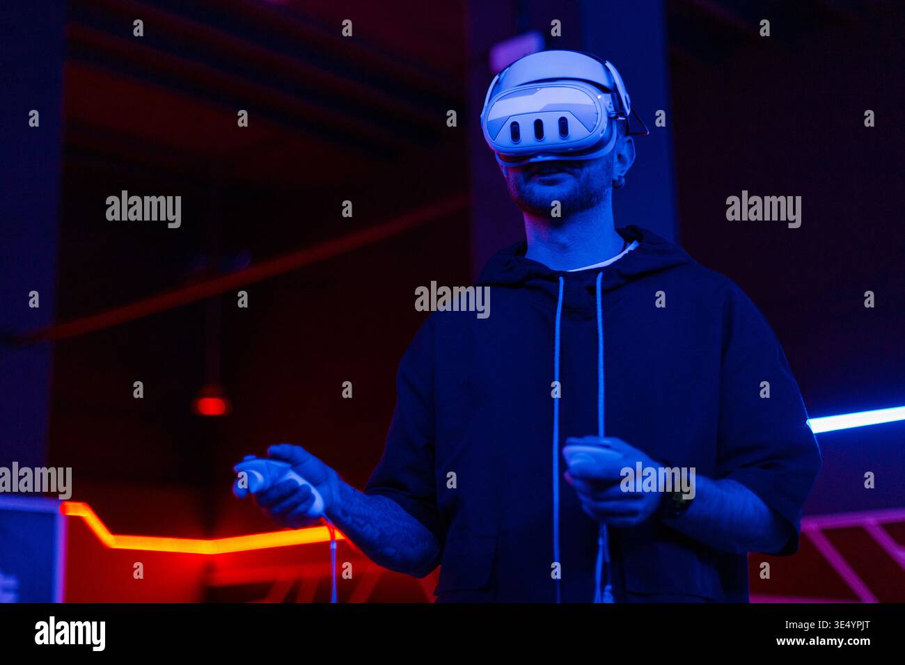 Ein Mann in einem schwarzen Hoodie mit VR-Brille und Joysticks in der Hand spielt Virtual-Reality-Spiele in einem dunklen Raum mit Neonlicht Stockfoto