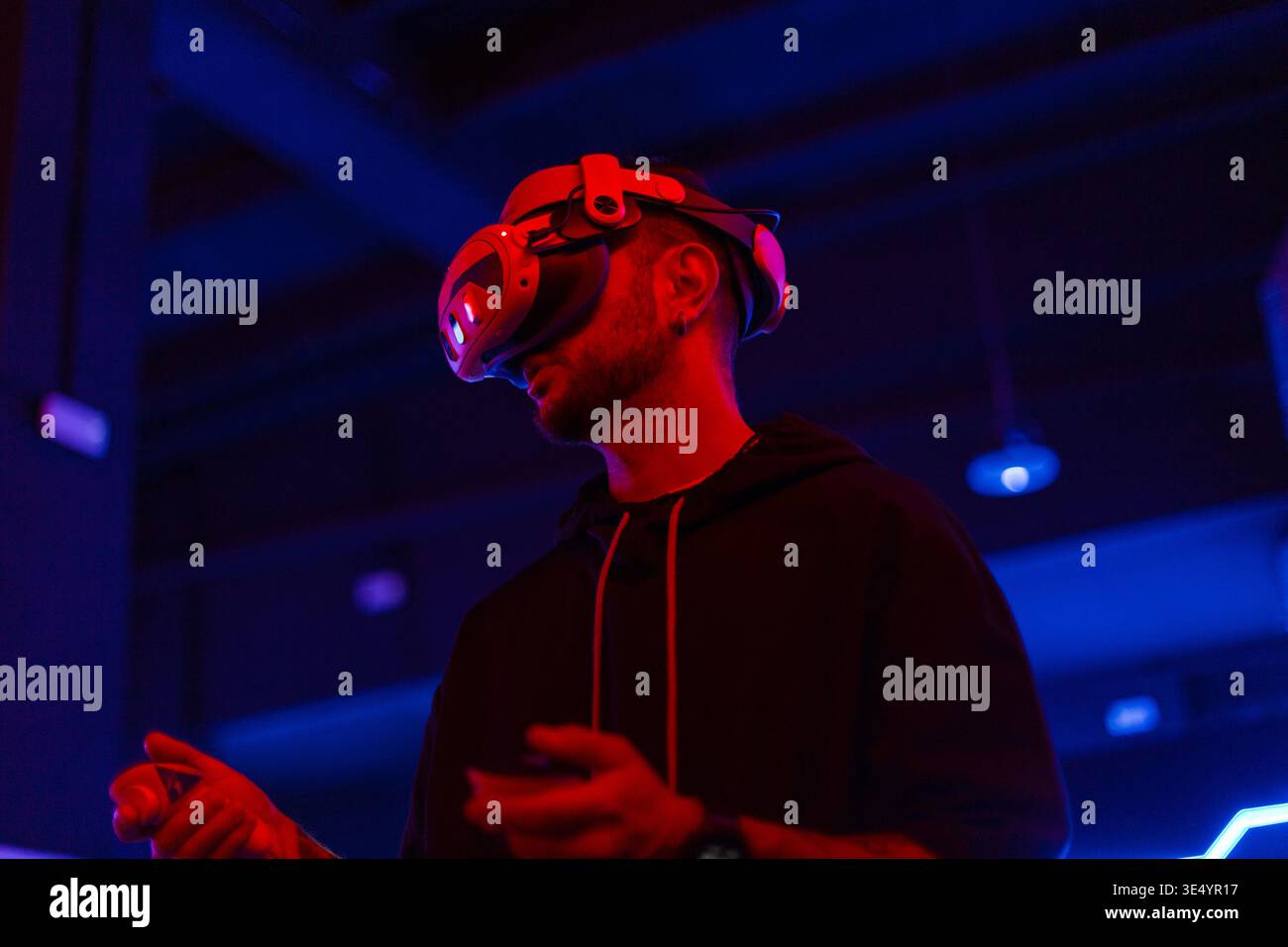 Cooler, gutaussehender professioneller Mann mit VR-Brille mit Joystick, der Virtual-Reality-Spiel in dunklem Hintergrund mit rotem und blauem Licht spielt Stockfoto