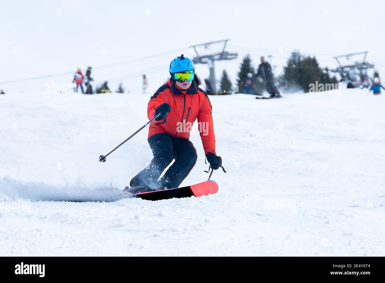 Blick auf einen Skifahrer in einer orangen Jacke, der eine schneebedeckte Piste hinabschneidet, mit Sesselbahnen in der Ferne, vor weißer Winterkulisse, Val-d Illiez, Wallis Stockfoto