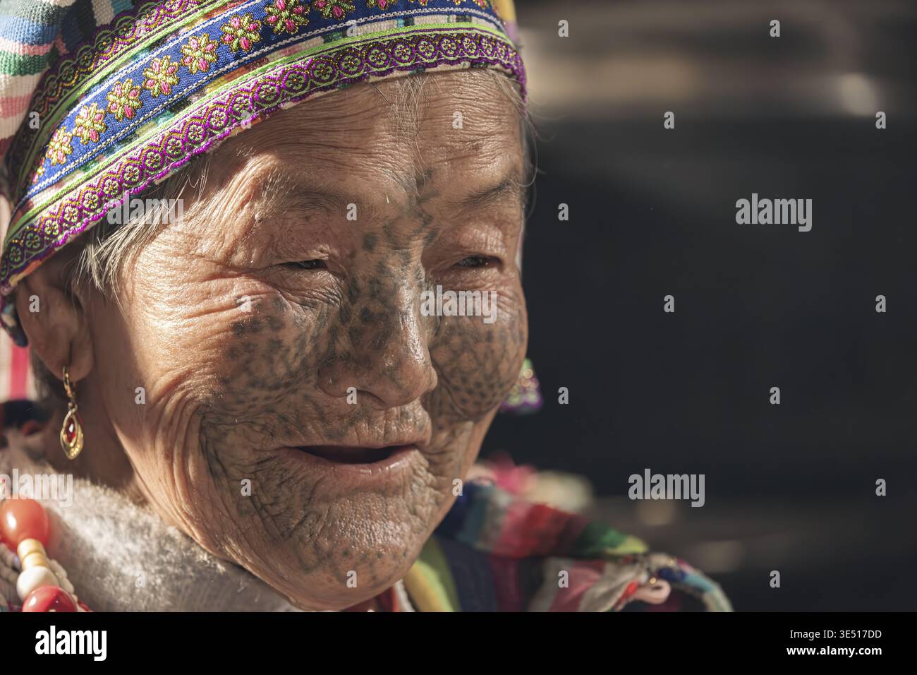 Dulong, China - 12. März 2026: Blick auf das Gesicht einer älteren Dulong-Frau mit traditionellen Tätowierungen, eingerahmt von einem lebendigen Kopfschmuck, ihr Lächeln ein Testamen Stockfoto