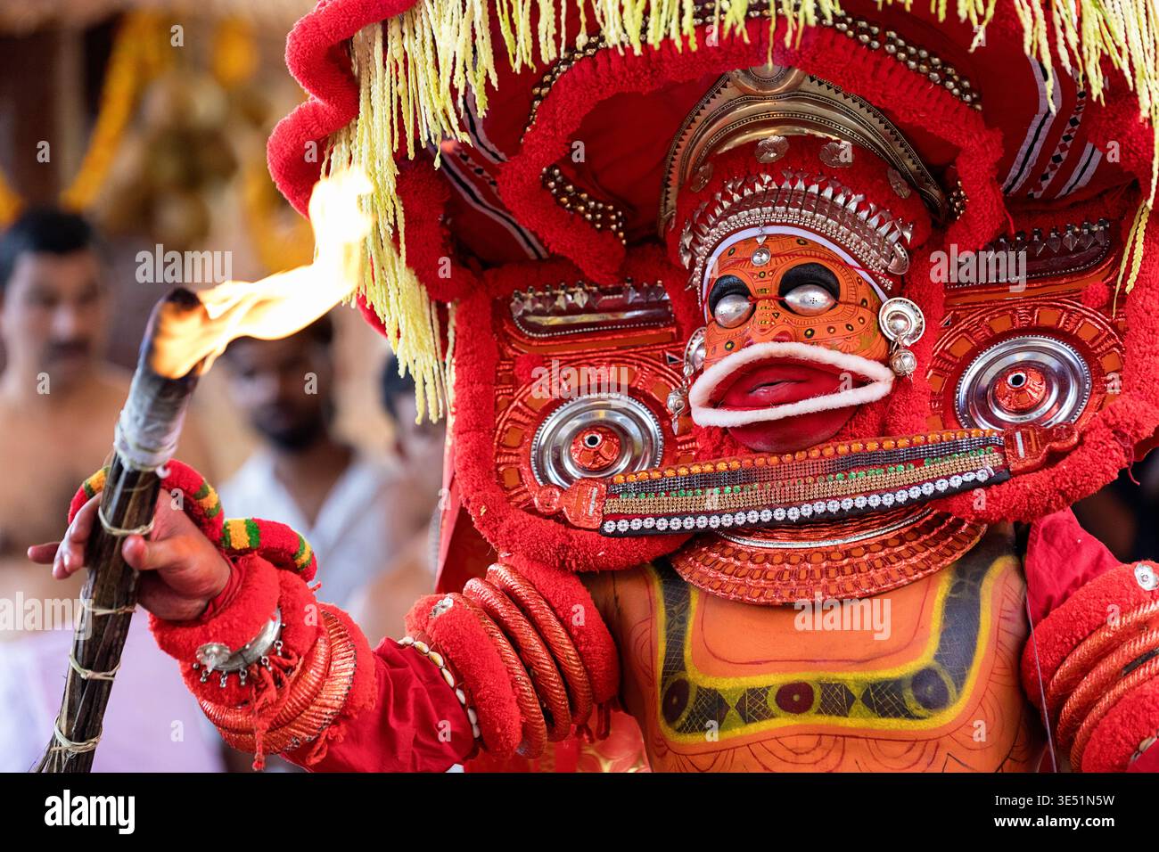 Theyyam-Darsteller in traditioneller roter Tracht und ausgeklügelter Gesichtsfarbe mit einer brennenden Fackel während eines rituellen Tanzes; Kannur, Kerala, Indien Stockfoto