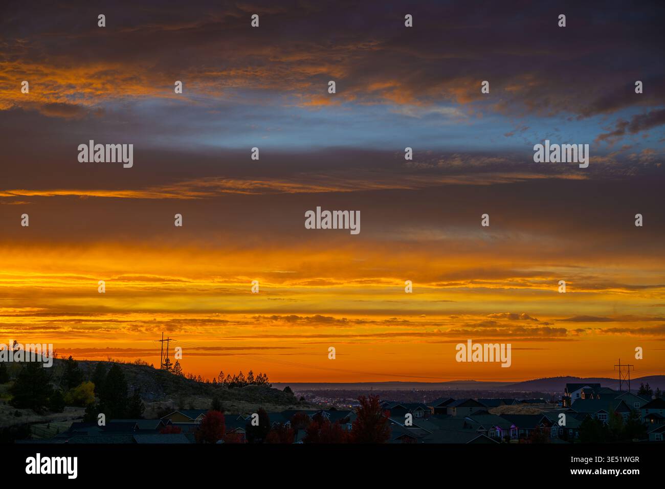 Hohe Hochspannungsleitungen verlaufen über eine bergige Vorstadtsiedlung mit Stadtlichtern in der Ferne unter einem farbenfrohen Sonnenuntergangshimmel. Stockfoto