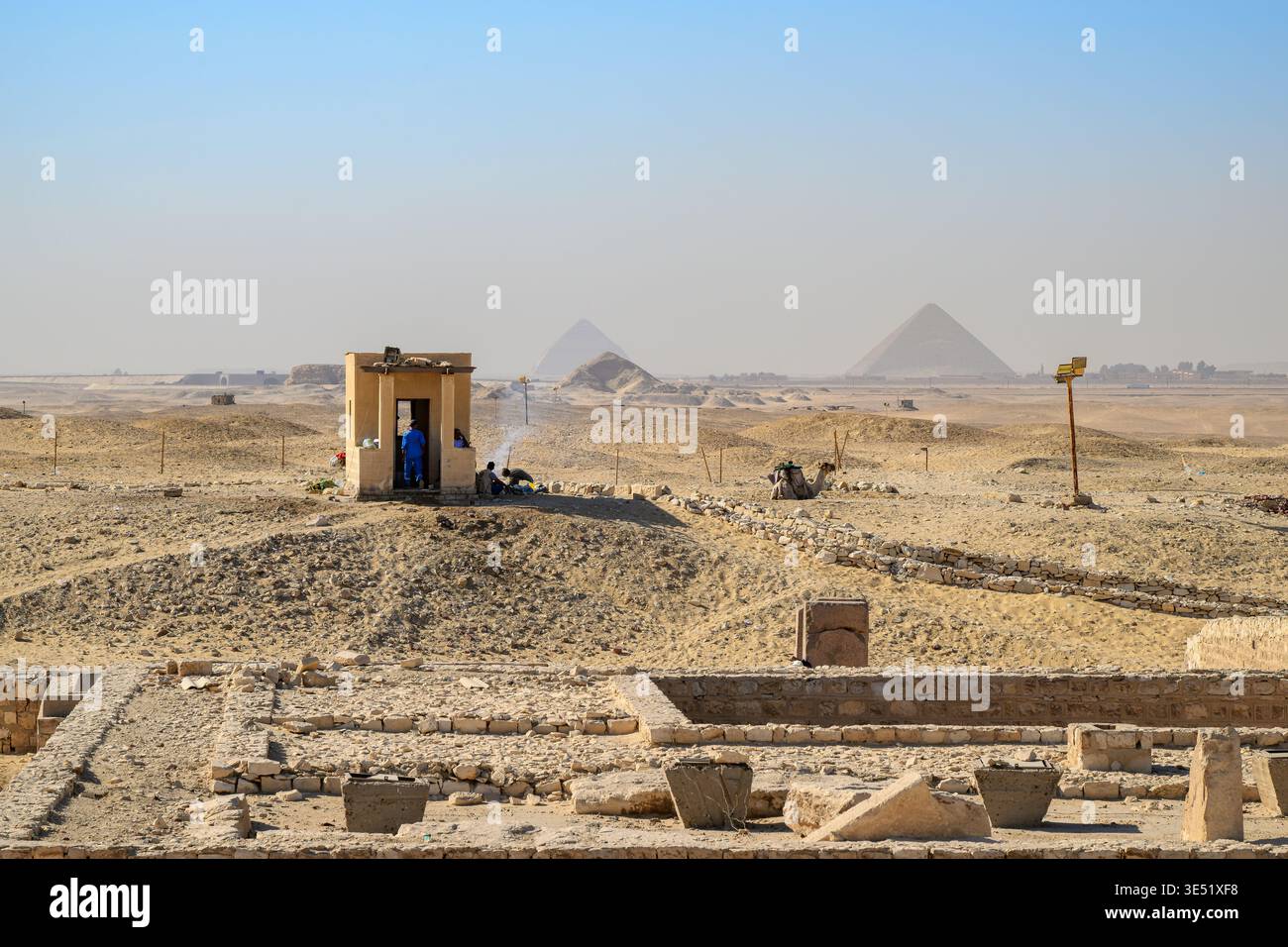 Die Ruinen der ägyptischen Nekropole von Sakkara auf dem Gizeh-Plateau mit Arbeitern, die eine Pause im Schatten machen, und der Komplex der Großen Pyramiden im Blick dahinter. Stockfoto
