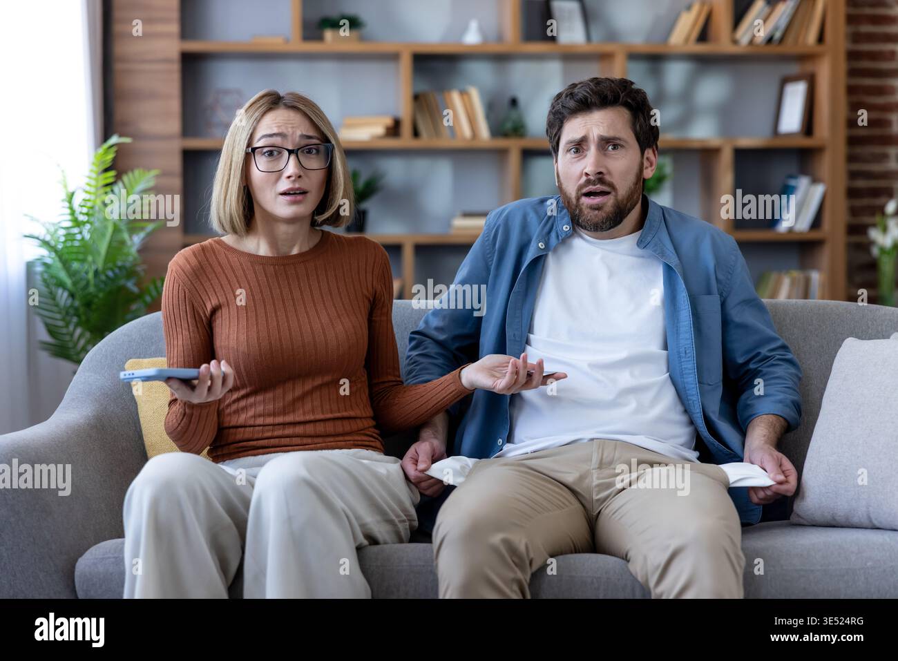 Frustriertes junges Paar, das zu Hause auf einem Sofa sitzt, finanzielle Schwierigkeiten hat und leere Taschen zeigt und sich über Konkurs und Geldprobleme gestresst fühlt Stockfoto