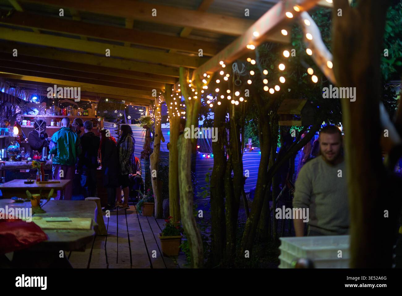 Ein abendliches geselliges Treffen im Freien in einem rustikalen Garten, beleuchtet von warmen Lichtern, die an Holzbalken und Bäumen hängen. Mehrere Personen Stockfoto