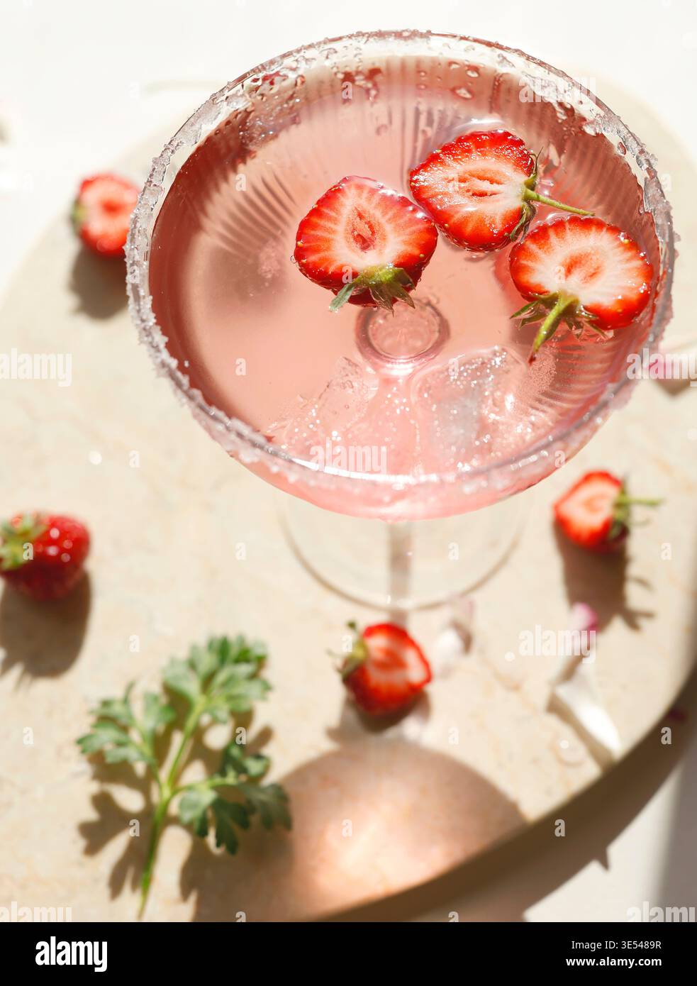 Rosafarbener Erdbeermargarita-Cocktail mit Zuckerrand auf Marmorschale. Frische halbierte Beeren, die in einem spritzigen Rosengetränk mit Eis und Sonnenlicht schwebt Stockfoto