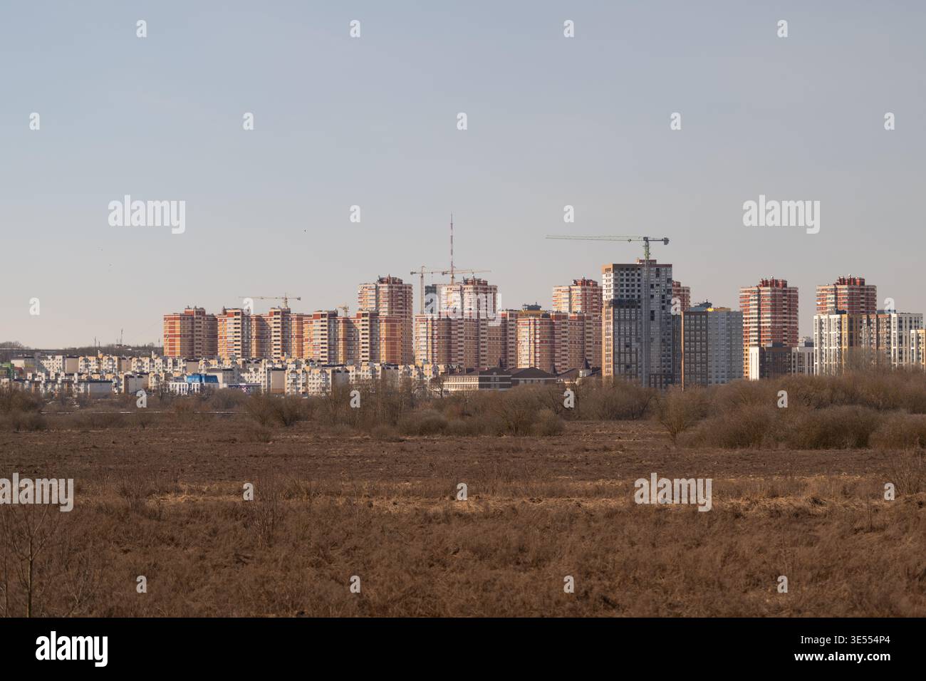 Ferne moderne Apartmentgebäude am Horizont über trockenem Grasland, Kräne und klarem Himmel, warmes Licht auf neuen Blöcken, Wohntürme mit Bauten Stockfoto