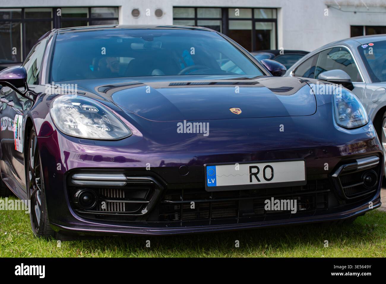 Valmiera, Lettland-26. April 2025-Dunkelviolette Porsche Panamera mit lettischem Kennzeichen, das bei einem Autoevent draußen auf Gras geparkt wurde, umgeben von anderen Lu Stockfoto