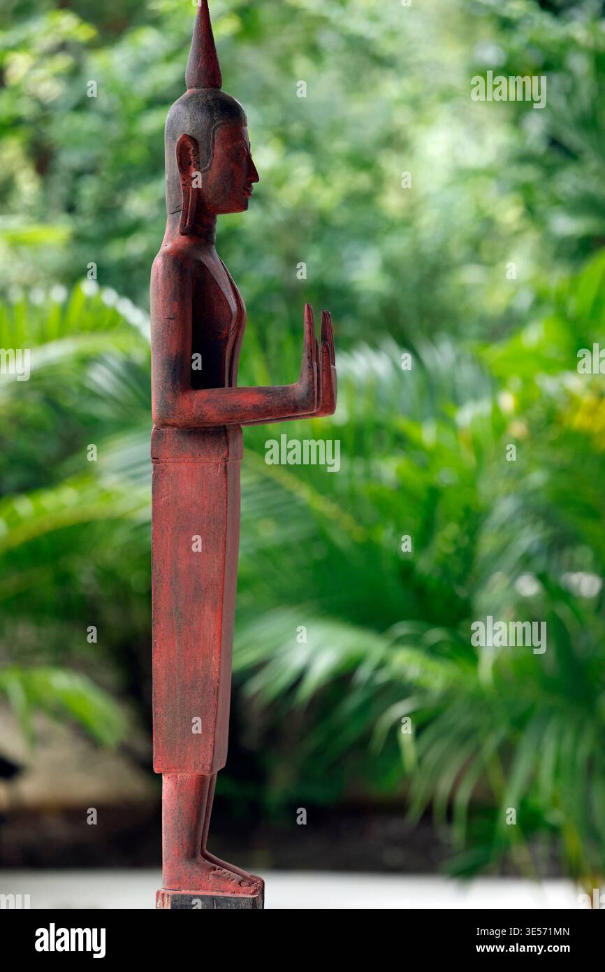 Antike rote Buddha-Figur aus Holz, aufrecht stehend mit erhobener Hand im üppigen grünen Garten. Siem Reap. Kambodscha. Stockfoto