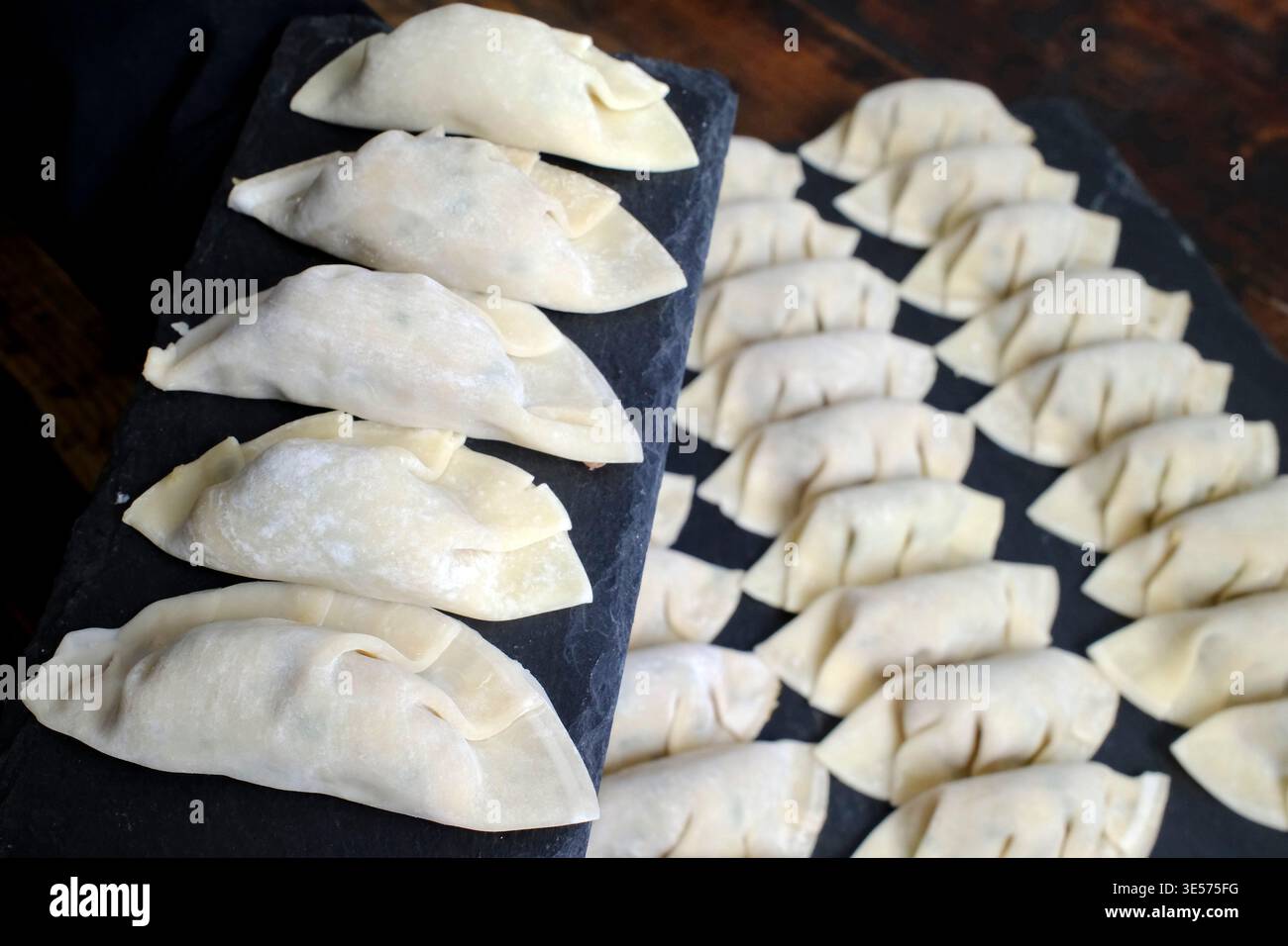 Ungekochte chinesische Teigtaschen mit kontrastierenden Falttechniken auf schwarzer Schieferoberfläche. Stockfoto