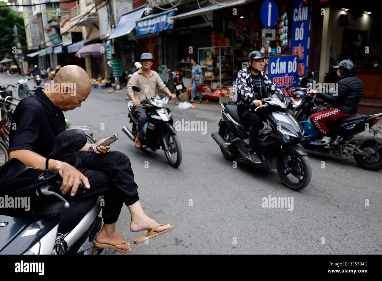 Mann, der das Telefon auf dem Motorrad durchblättert, während der Verkehr durch den lebhaften vietnamesischen Straßenmarkt fließt. Haiphong. Vietnam. Stockfoto