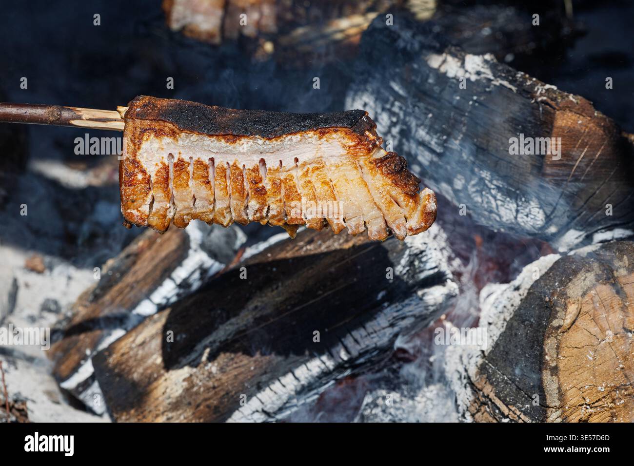Knusprige Speckstreifen, die über einem offenen Lagerfeuer gegrillt werden, erzeugen Rauch und goldverkohlte Kanten. Rustikale Outdoor-Kochszene, die Wärme weckt, Stockfoto