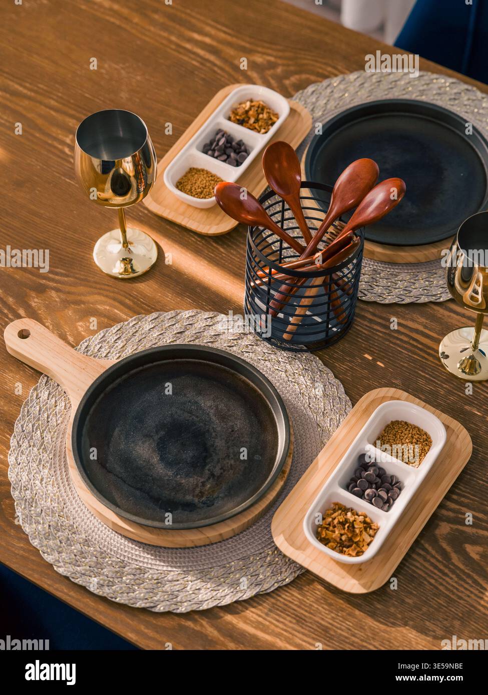 Eine symmetrische Anordnung von Küchenutensilien, einschließlich Eisenpfannen und goldenen Bechern, steht auf einem braunen Tisch bei weichem, diffusem Tageslicht. Stockfoto