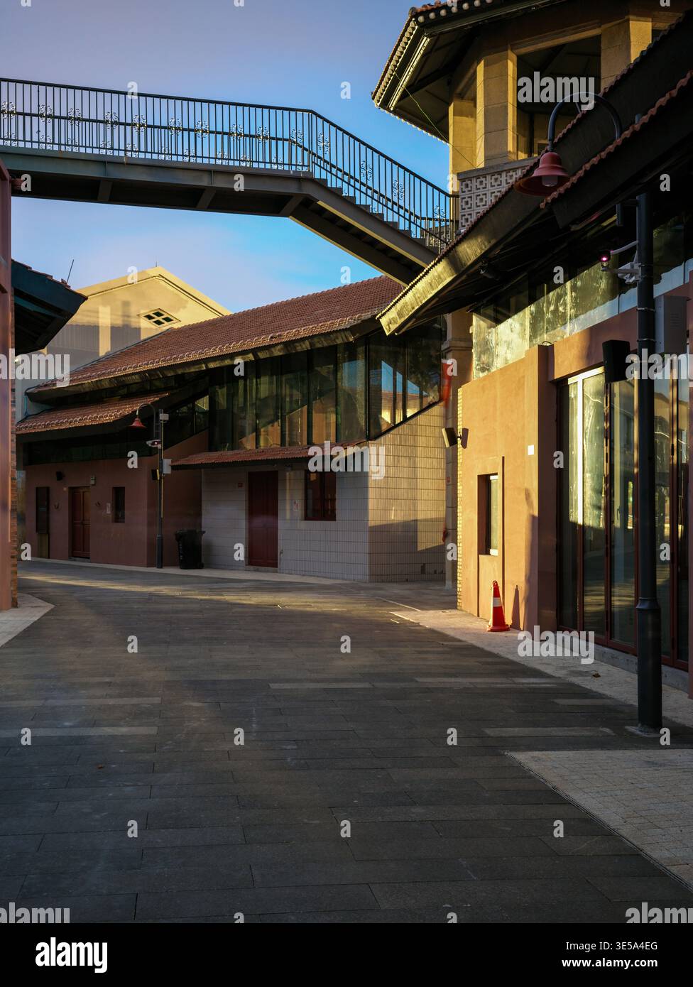 Leere gepflasterte Fußgängerzone mit einem erhöhten Metallsteg, der zwei renovierte Industriegebäude mit starkem Sonnenlicht und tiefen Schatten überbrückt. Stockfoto