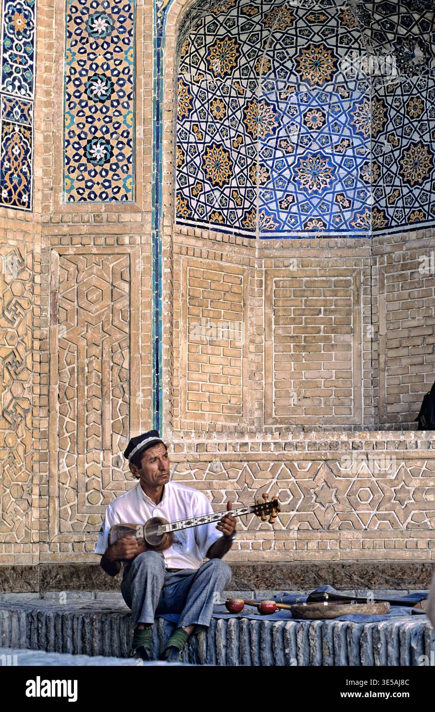 Ein Musiker sitzt an einer wunderschönen Mauer und spielt ein Streichinstrument in Buchara, Usbekistan. Er ist umgeben von historischen Mustern und Fliesen. Stockfoto
