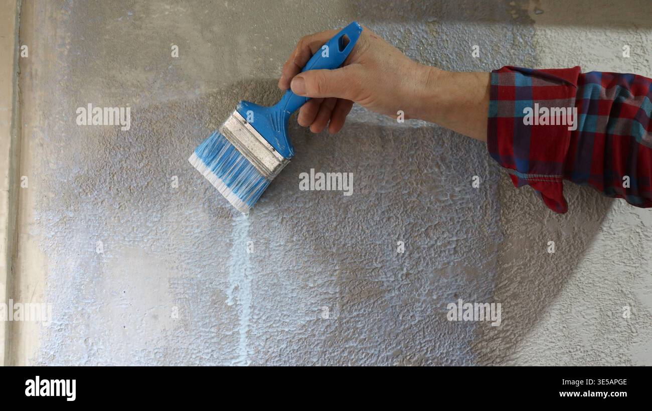 Nahaufnahme eines Mannes in kariertem Hemd mit blauem Pinsel, der flüssige Grundierung auf strukturierte graue Putzwand und Grundierwand des Hauses aufträgt Stockfoto
