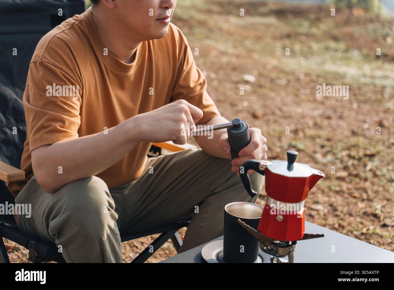 Männlicher Reisender sitzt in einem Klappstuhl mit manueller Mahlmaschine, um frischen Espresso und eine Kaffeemaschine auf dem Bergcamping zuzubereiten Stockfoto