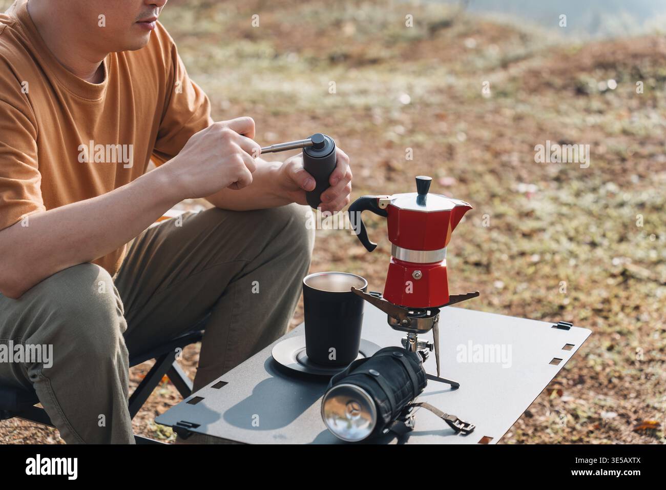 Männlicher Reisender sitzt in einem Klappstuhl mit manueller Mahlmaschine, um frischen Espresso und eine Kaffeemaschine auf dem Bergcamping zuzubereiten Stockfoto