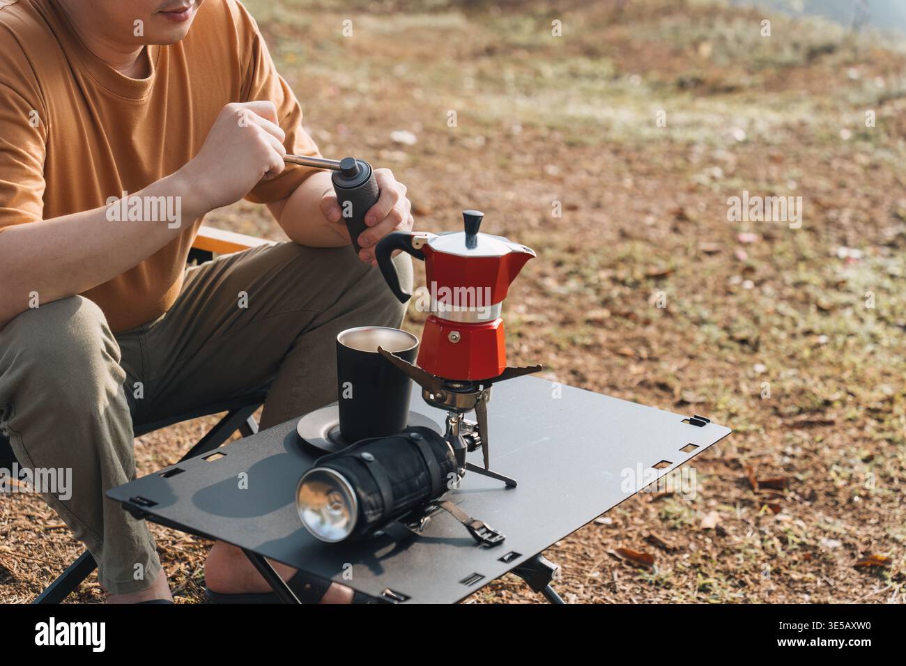 Männlicher Reisender sitzt in einem Klappstuhl mit manueller Mahlmaschine, um frischen Espresso und eine Kaffeemaschine auf dem Bergcamping zuzubereiten Stockfoto
