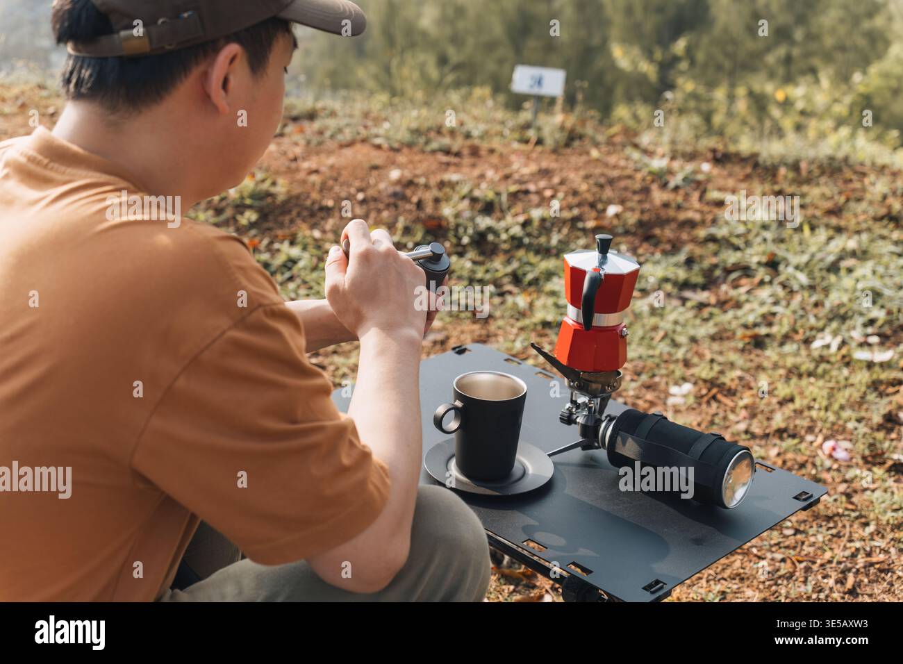 Männlicher Reisender sitzt in einem Klappstuhl mit manueller Mahlmaschine, um frischen Espresso und eine Kaffeemaschine auf dem Bergcamping zuzubereiten Stockfoto