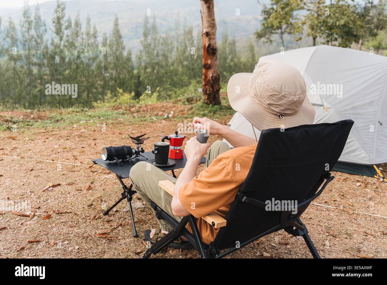 Männlicher Reisender sitzt in einem Klappstuhl mit manueller Mahlmaschine, um frischen Espresso und eine Kaffeemaschine auf dem Bergcamping zuzubereiten Stockfoto