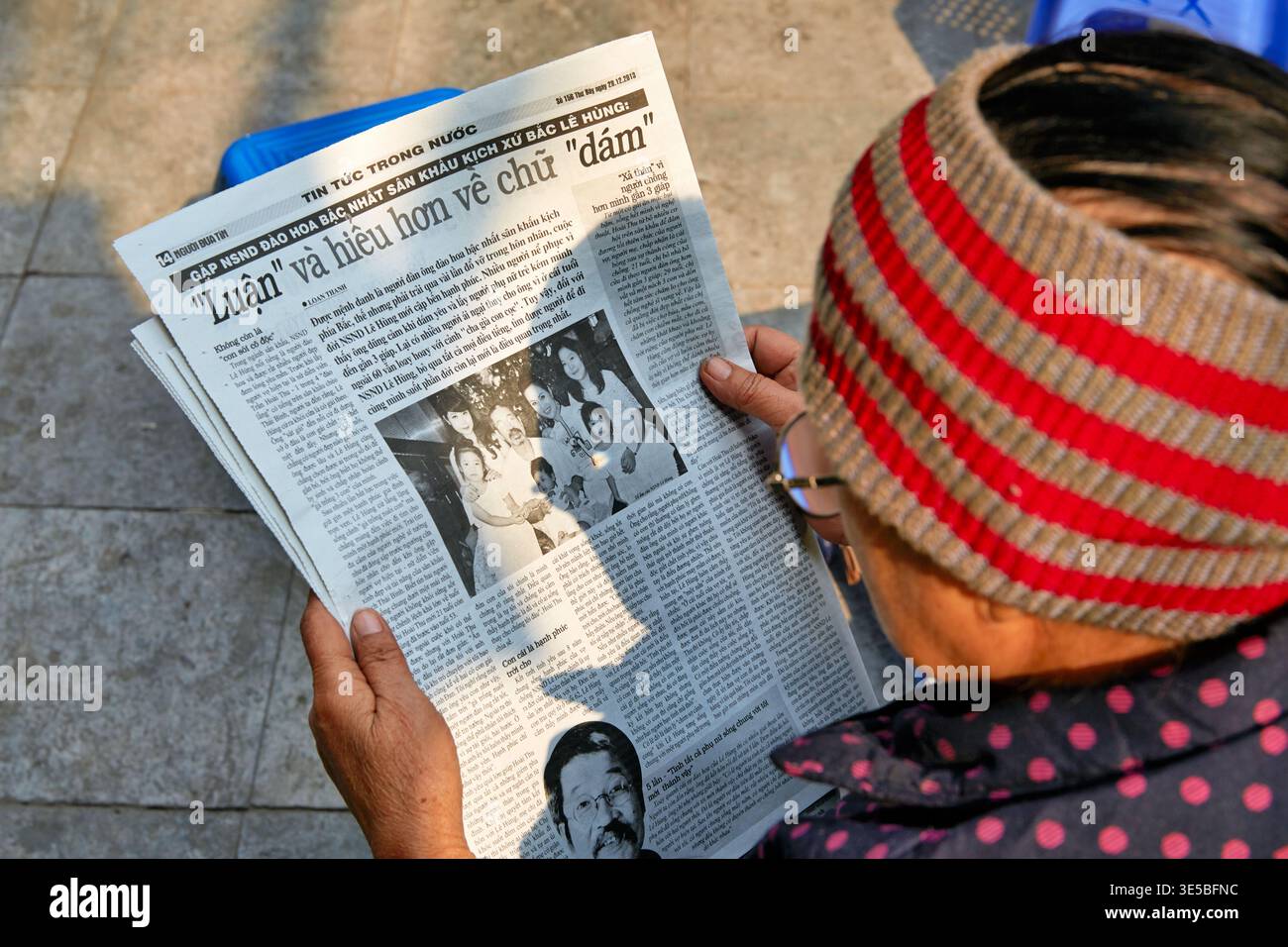 Eine ältere Person hält eine Zeitung, während sie draußen in Hanoi sitzt. Die Szene fängt das tägliche Leben in Vietnam tagsüber ein. Stockfoto