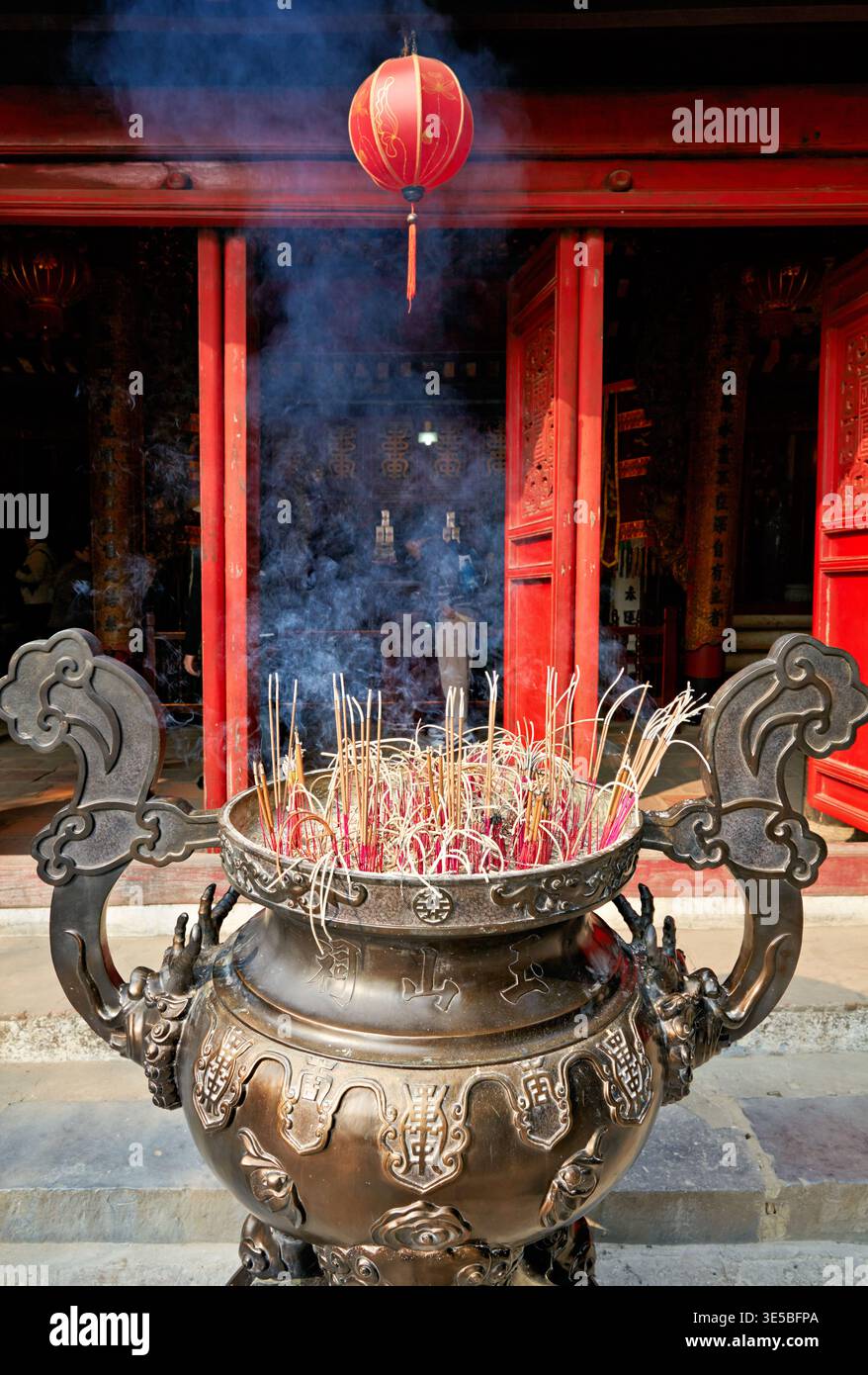 Besucher beobachten, wie der Rauch aus einem Räucherbrenner vor einem Tempel in Hanoi, Vietnam, während eines geschäftigen Tages voller kultureller Aktivitäten aufsteigt. Stockfoto