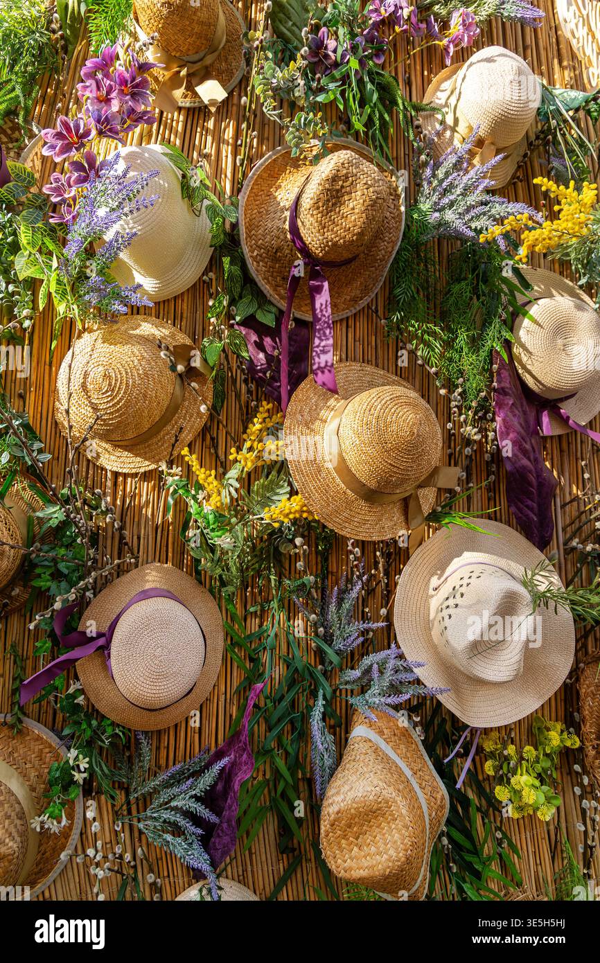 Strohhüte und Blumen auf Bambuswand. Dekorative Präsentation mit gewebten Strohhüten, Bändern und bunten Blumen, die auf einer Bambuswand angeordnet sind, sorgen für eine fröhliche Stimmung Stockfoto