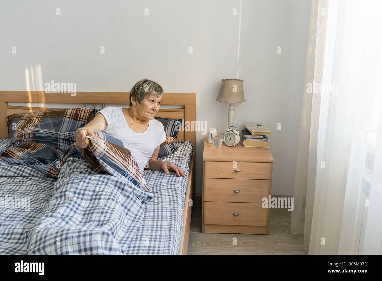 Eine ältere Frau sitzt im Bett, blickt zum Fenster und bereitet sich auf den Start in den Tag vor. Morgenlicht in das Schlafzimmer. Das wirkliche Leben eines älteren Menschen Stockfoto
