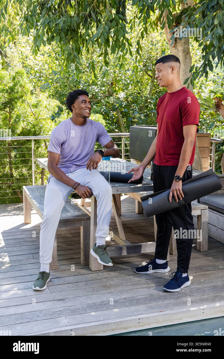 Verschiedene männliche Freunde, die auf einer Bank an einem Tisch auf dem Hinterhof sitzen, mit gerollten Yogamatten Stockfoto