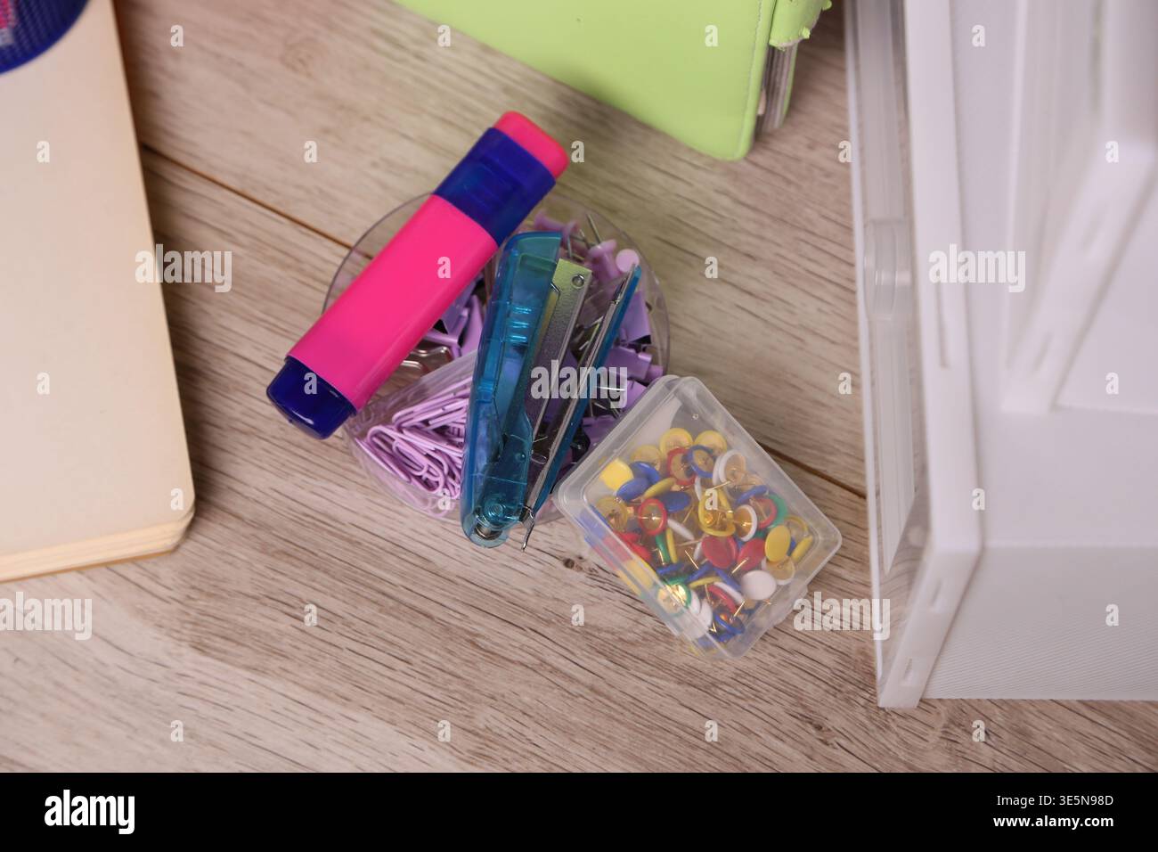 Eine Sammlung von Büromaterial liegt auf einem Holztisch. Es gibt Büroklammern, einen Textmarker und eine Schachtel mit Reißzwecken. Ein Notizbuch und ein grüner Ordner befinden sich in der Nähe und schaffen einen Arbeitsbereich Stockfoto