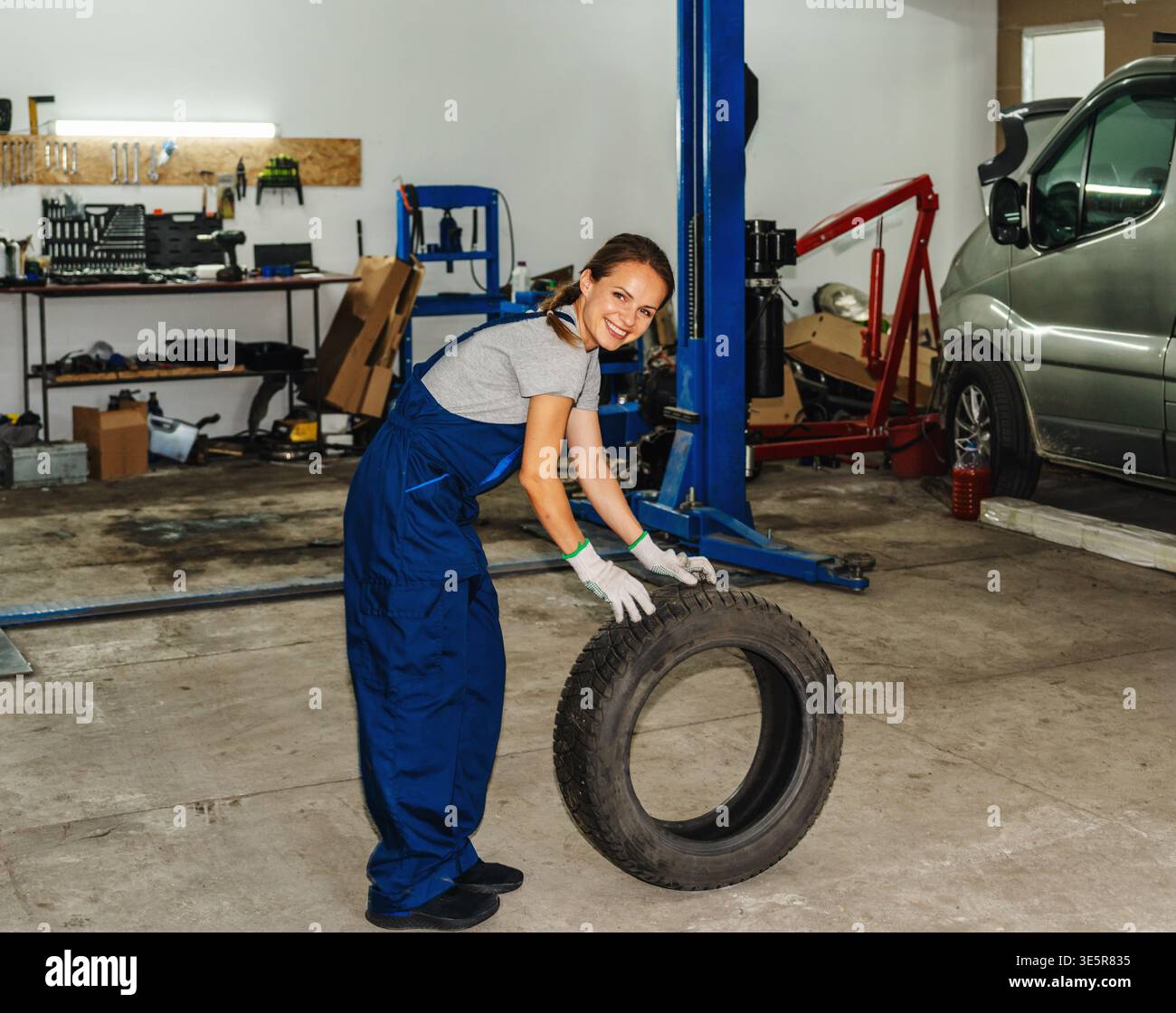 Die volle Länge einer lächelnden Mechanikerin, die in einer professionellen Autowerkstatt einen Autoreifen rollte. Stockfoto