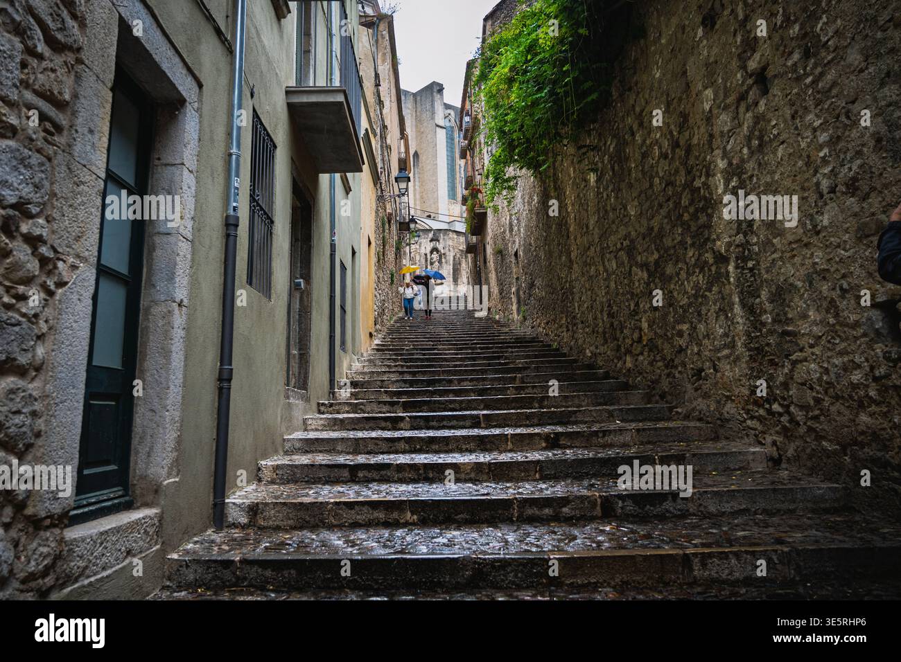Regnerische Steintreppen in der historischen Altstadt von Girona, Spanien, mit Menschen, die unter Sonnenschirmen laufen. Nasse Kopfsteinpflaster und eine enge mittelalterliche Straße schaffen einen Stockfoto