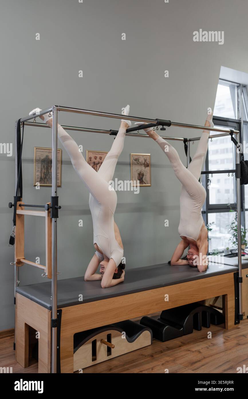 Das Pilates-Duo balanciert in umgedrehter Beinhaltung auf Reformertrainern im grauen Trainingsstudio zusammen. Umgedreht, Duo, Pilates, Reformer, Beinhaltung. Stockfoto