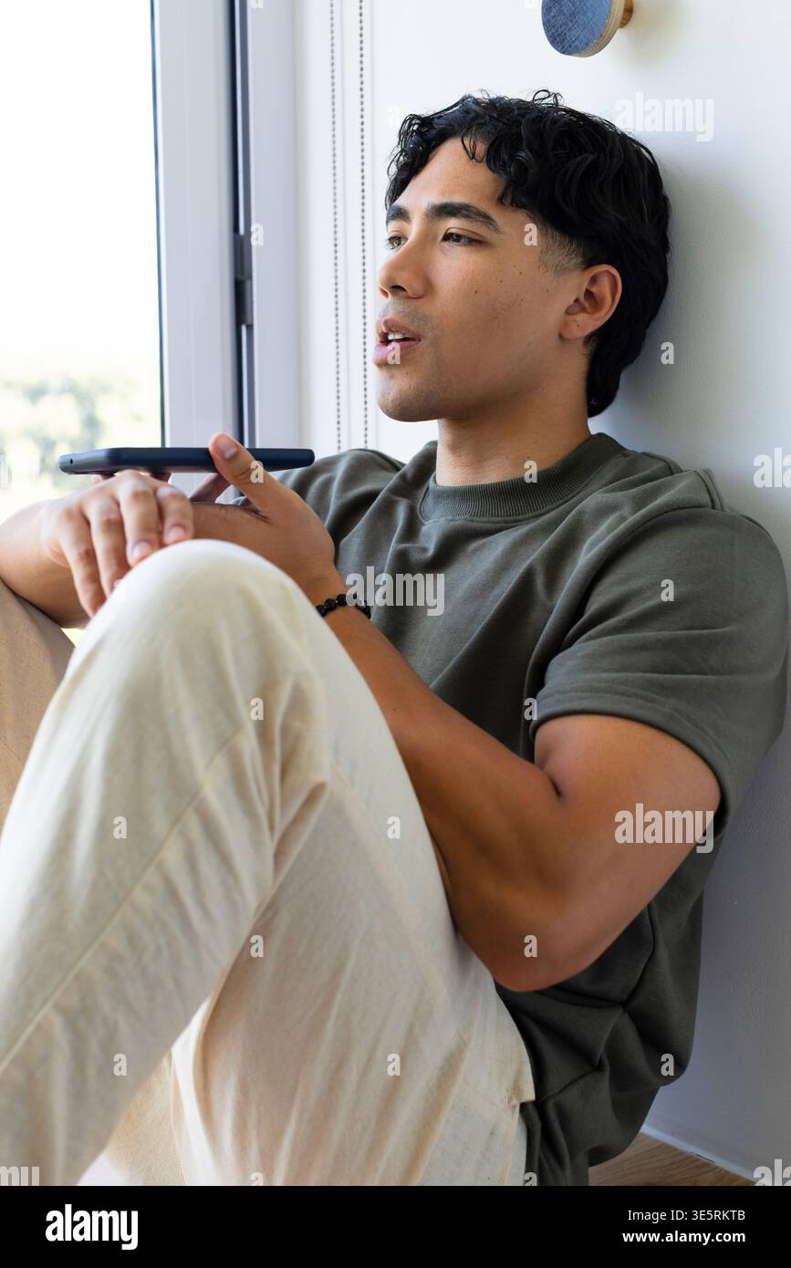 Asiatischer Mann, der an einem hohen Fenster sitzt, das Telefon waagerecht in der Nähe des Mundes hält und ein schwarzes Perlenarmband zeigt Stockfoto