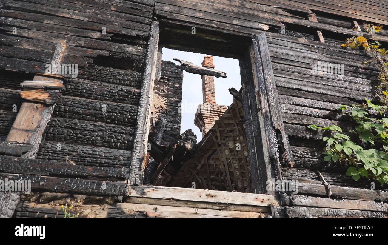 Verbrannte hölzerne Hauswand mit einem leeren Fensterrahmen und einem entfernten Ziegelkamin Stockfoto