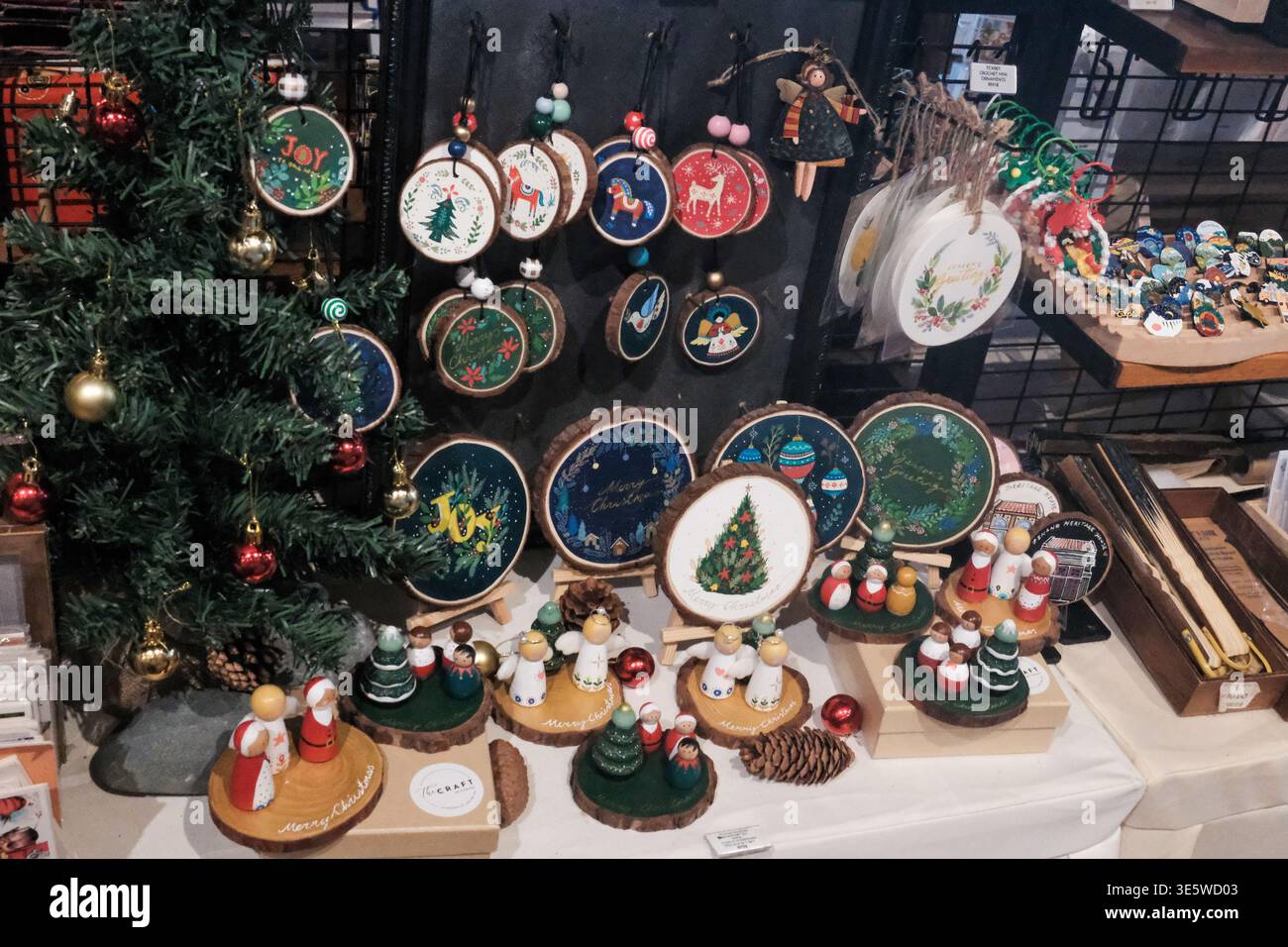 Festliche Ornamente zieren einen Weihnachtsmarkt-Stand. Kreisförmige Dekorationen hängen und sitzen auf Regalen. Miniaturbäume und Figuren schmücken das Holztablett Stockfoto