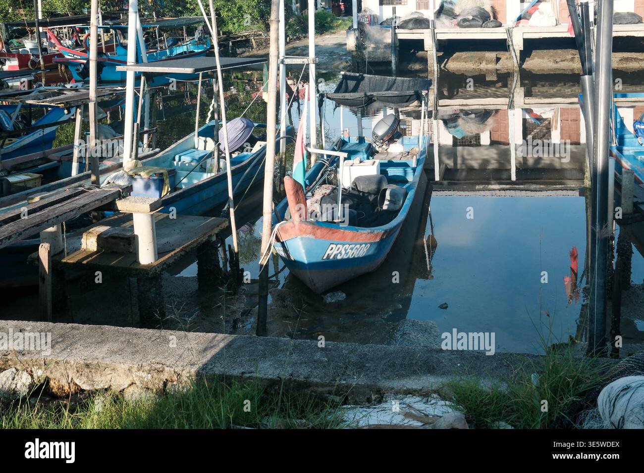 Das Boot ruht am hölzernen Dock neben ruhigem Wasser. Metallstangen stehen hoch neben dem Schiff. Graskante grenzt an den Rand des Docks. Schatten werden sanft über die Oberfläche geworfen Stockfoto
