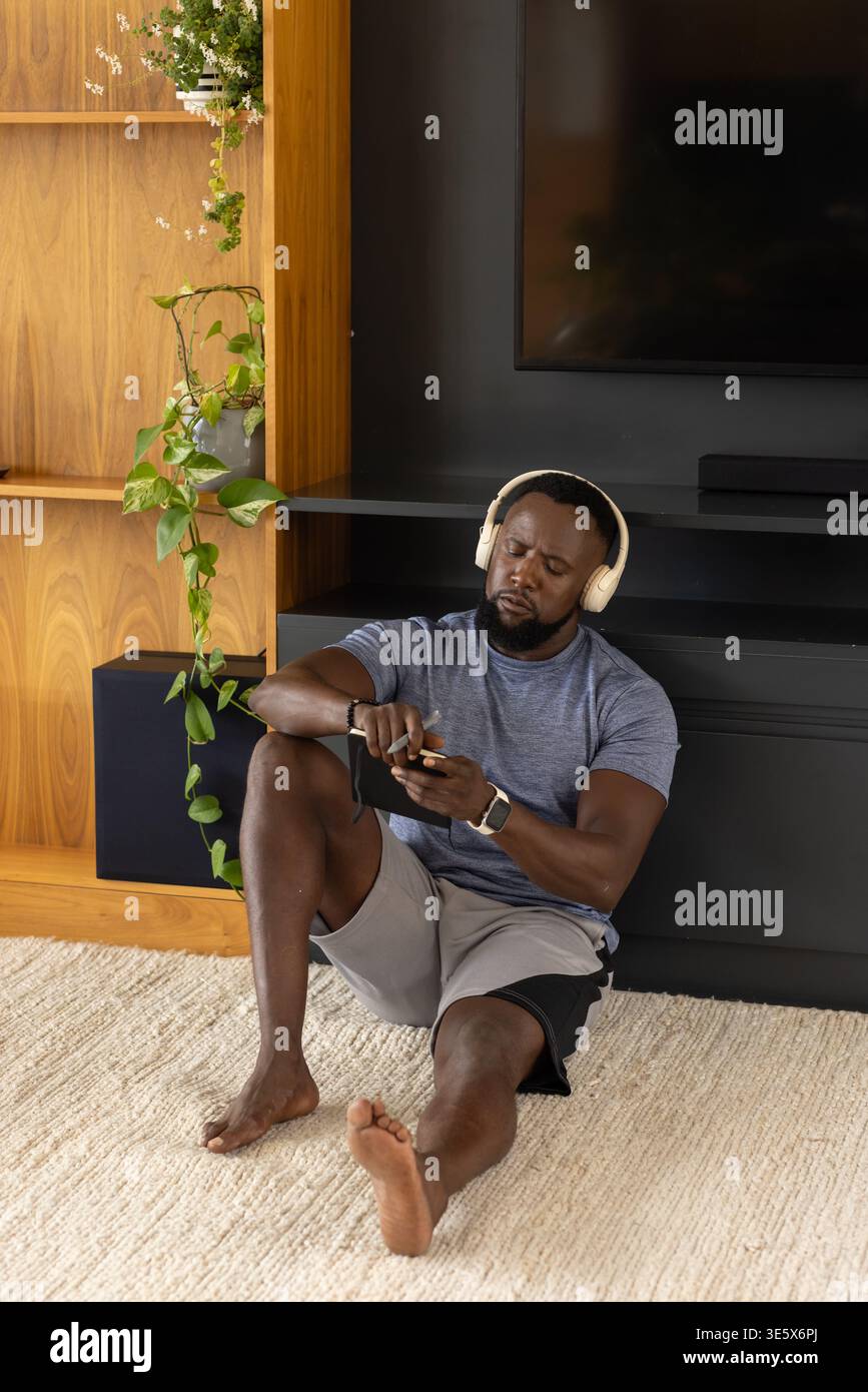 Erwachsener Mann sitzt auf weichem Teppich im Wohnzimmer, trägt Sportbekleidung und Kopfhörer, hält Telefon Stockfoto