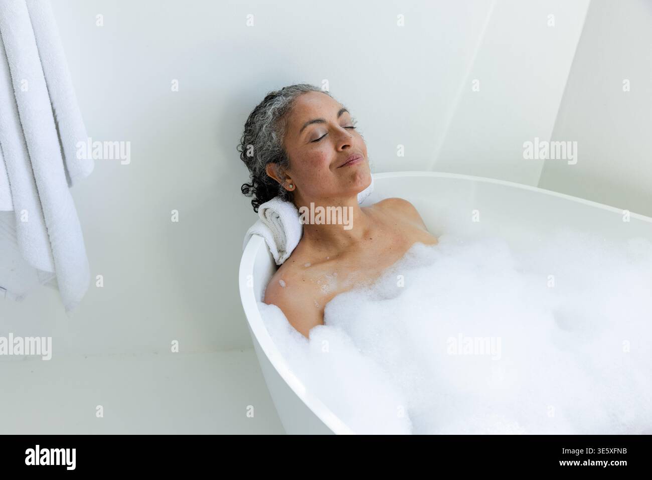 Ältere afroamerikanische Frau, die sich in einer freistehenden Wanne zurücklehnt, sich zwischen Seifenblasen entspannt, Handtuchkopfstütze Stockfoto