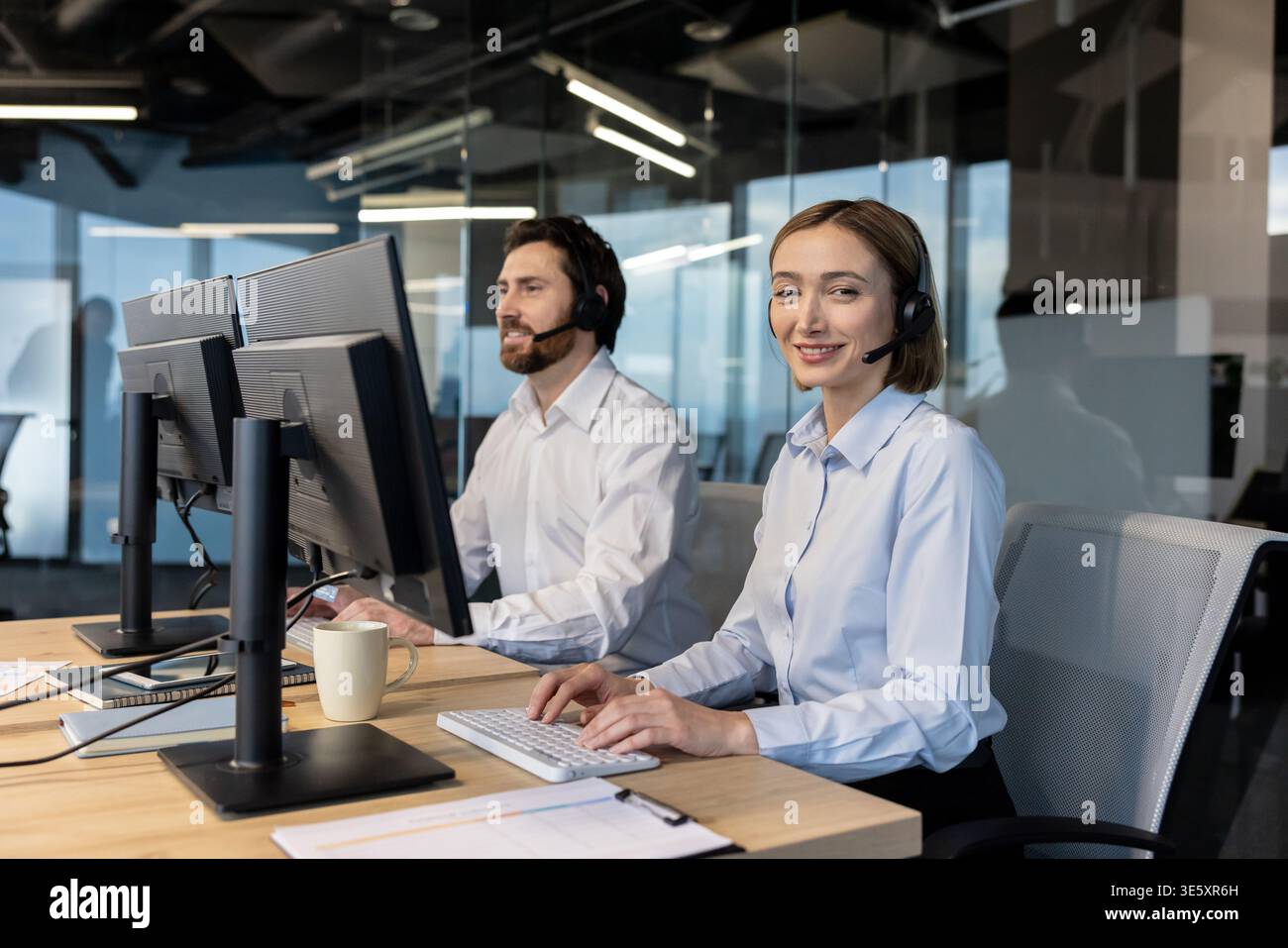 Lächelnde Frau, die beim Tippen am Computer ein Headset trägt, um einen Kollegen in einem modernen Kundenservice- oder Helpdesk-Umfeld freundlich zu unterstützen Stockfoto