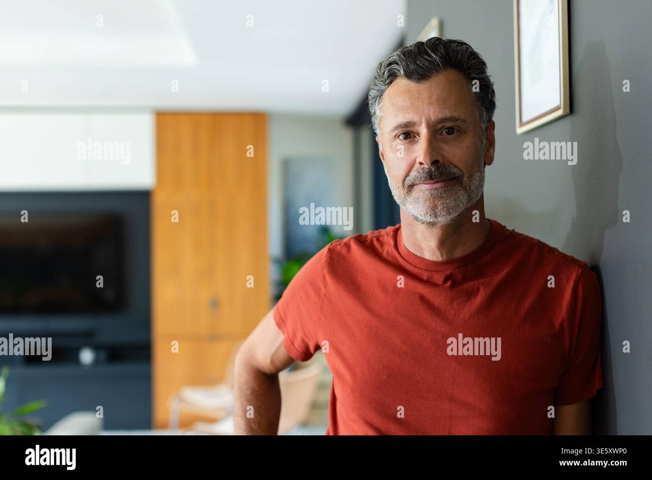Reifer Mann, der sich zu Hause in der Nähe des Fernsehens an die graue Wand lehnt, mit rotem T-Shirt, Kopierraum Stockfoto